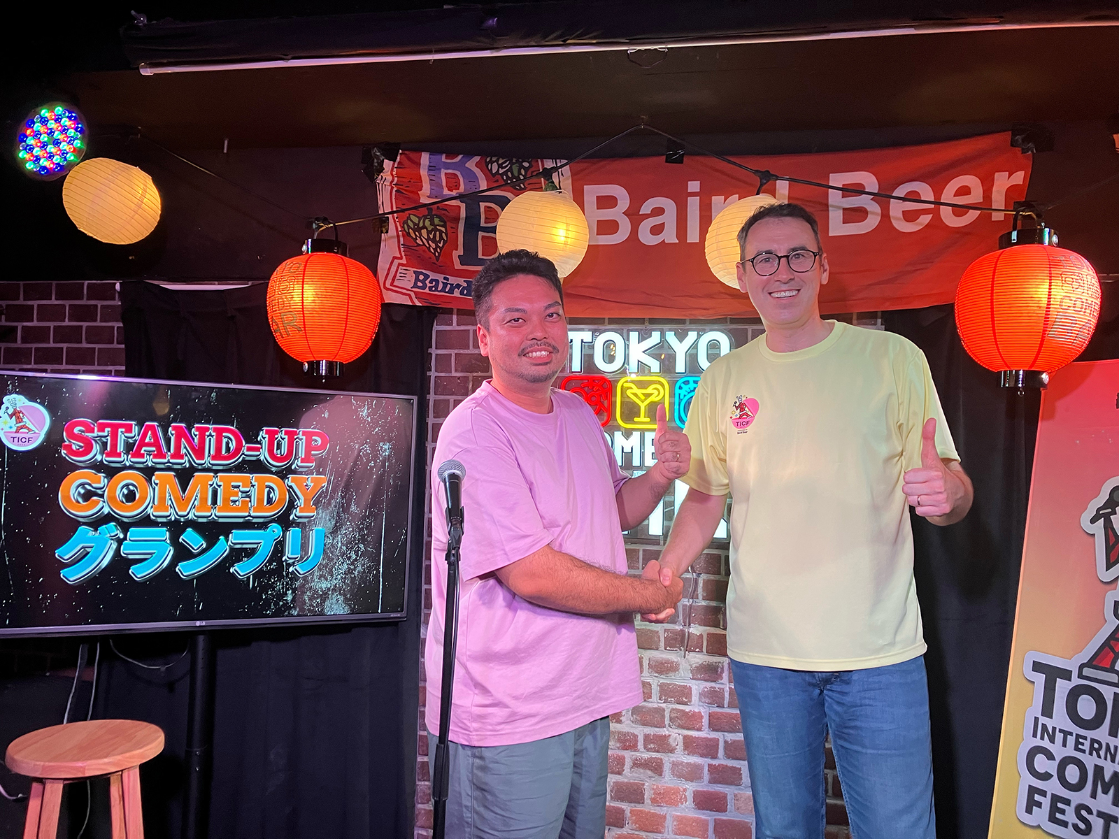 STAND-UP COMEDYグランプリの表示の横に立つ萱野公裕と男性